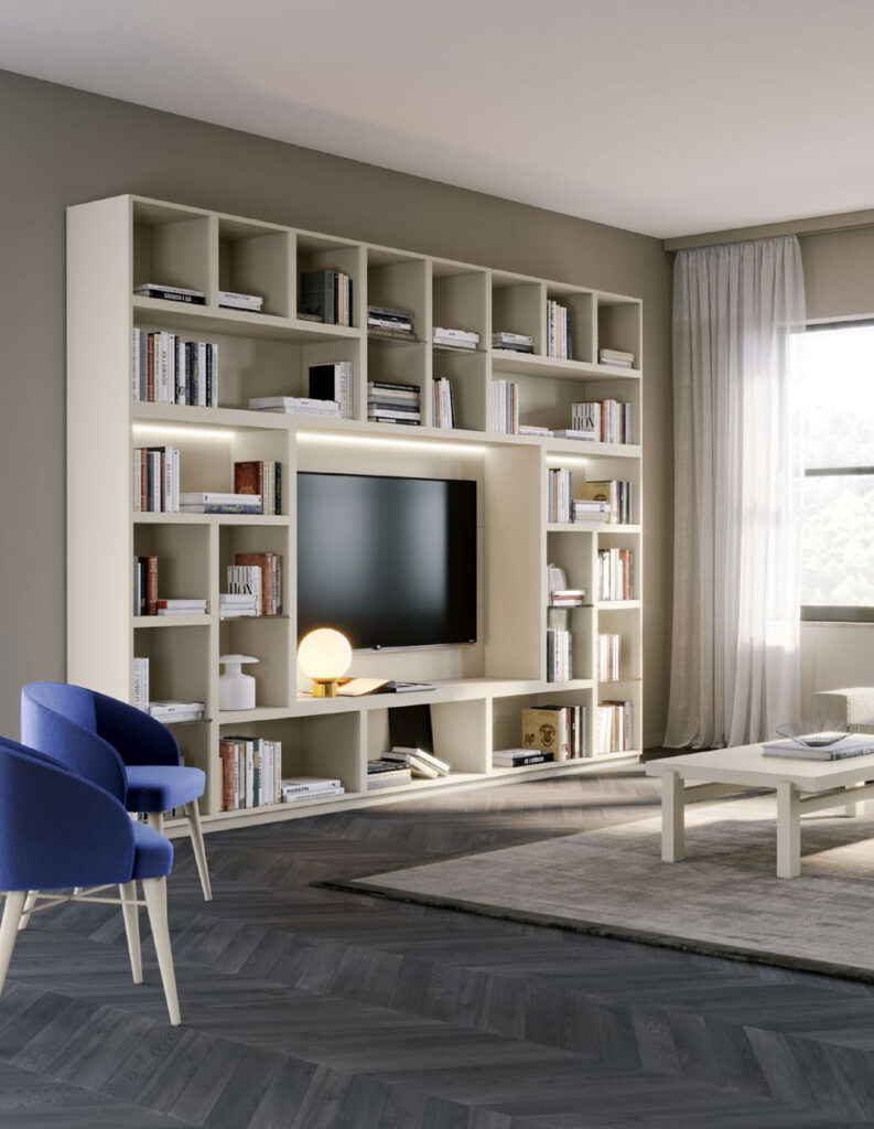 Soluzione d'arredo composta da mobili componibili per il soggiorno color bianco panna, composta elementi libreria aperti a tutta parete, con spazio centrale per la televisione. Dalla linea Quattrolati di Benigni Mobili. Prospettiva laterale. Soggiorno modulare.